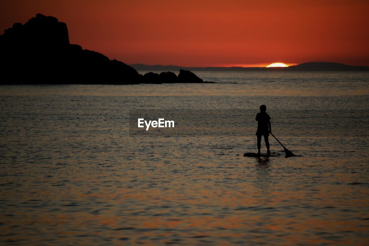 Silhouette person paddleboarding on sea at sunset