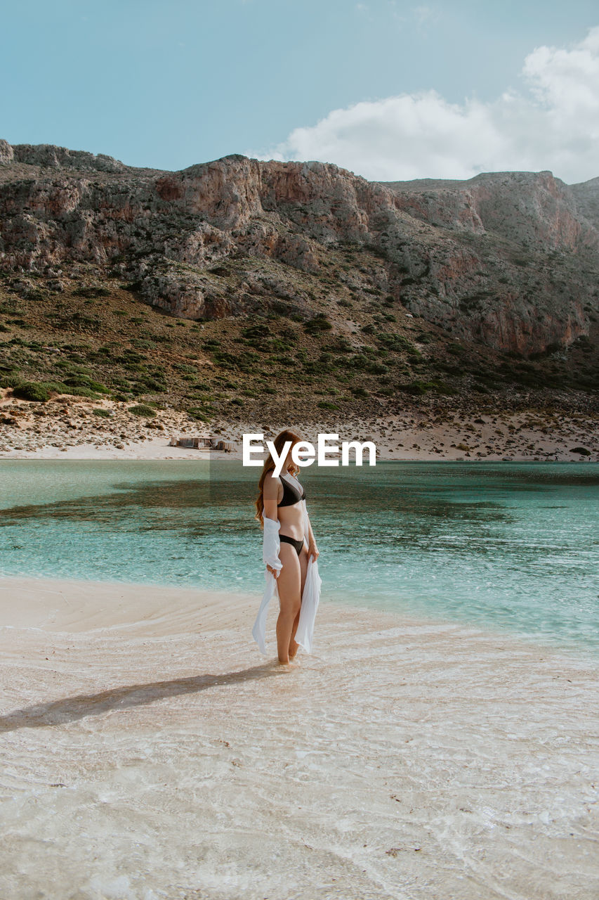 Full length slim female in black swimsuit standing on sandy balos beach against rocky cliff and looking away on clear sunny weather