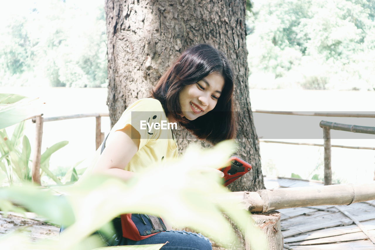 Smiling young woman using smart phone while sitting by tree trunk at park