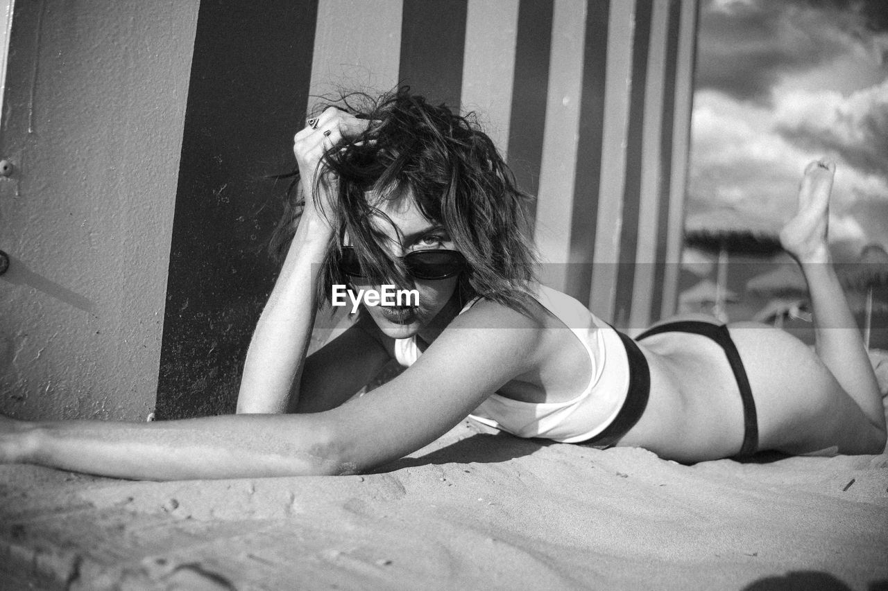 Portrait of young woman wearing bikini and sunglasses lying on sand at beach 