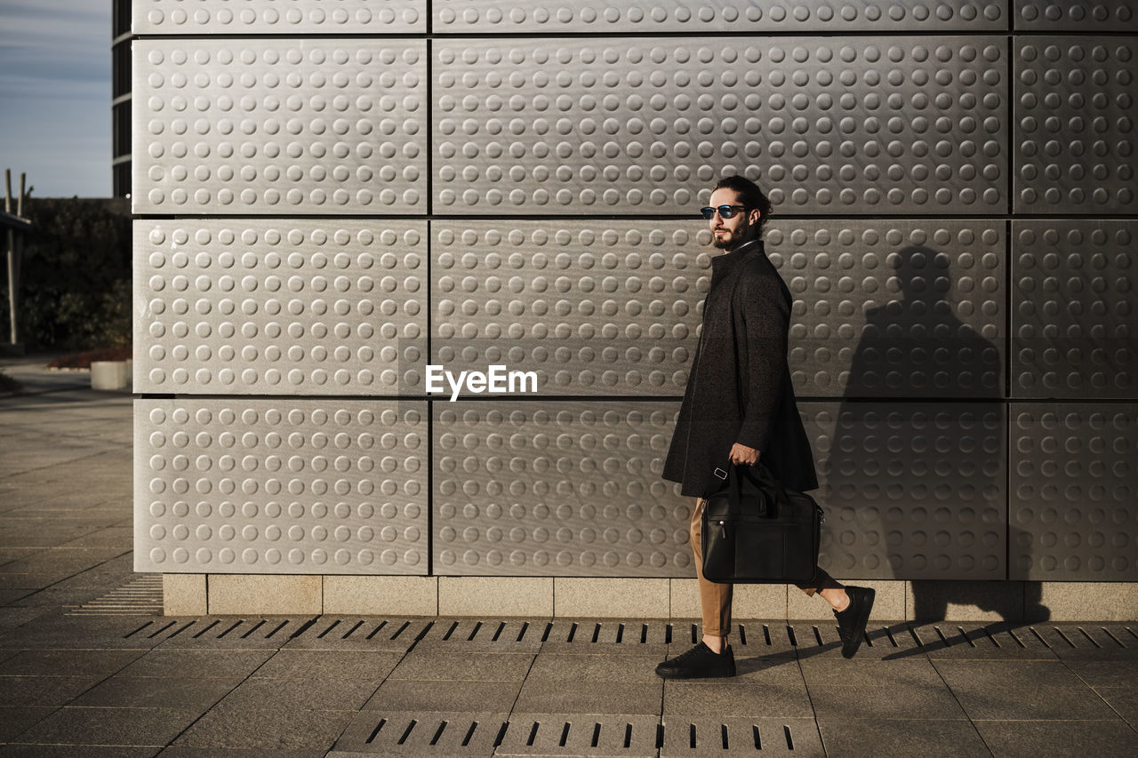 Businessman wearing jacket carrying briefcase while walking against silver colored wall