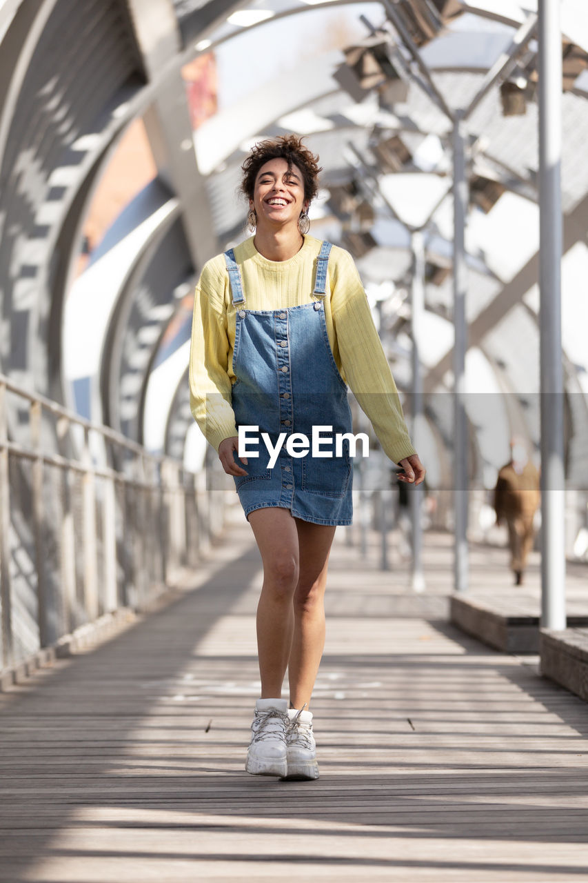 Low angle full body of happy young hispanic female with curly hair wearing denim overall dress with yellow sweatshirt and sneakers walking on enclosed footbridge in city