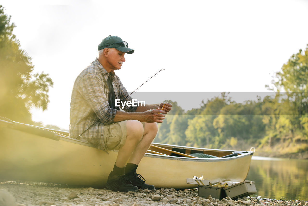 Senior man adjusting fishing tackle while sitting on boat at lakeshore against clear sky