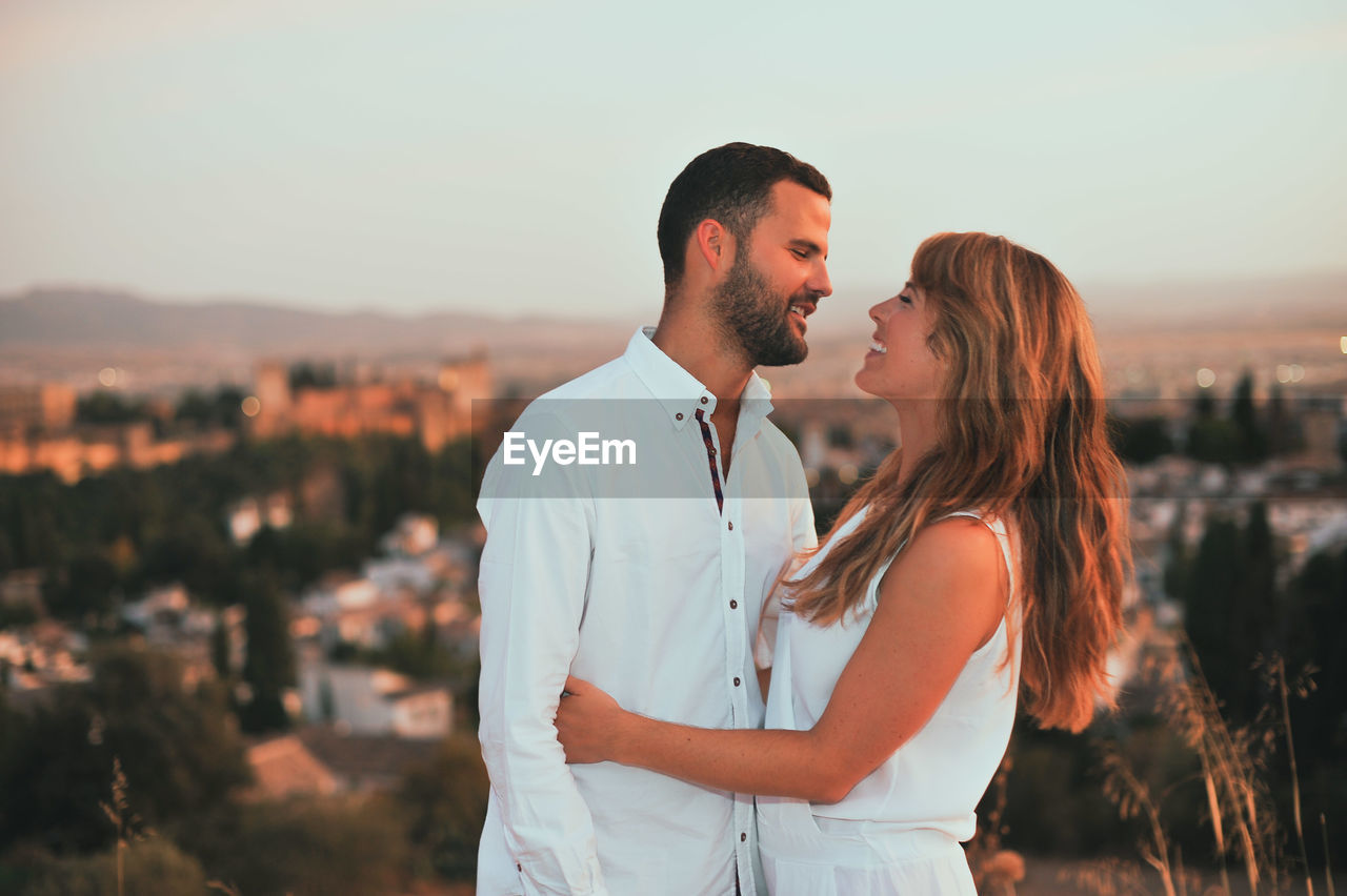 Happy romantic couple embracing against sky during sunset