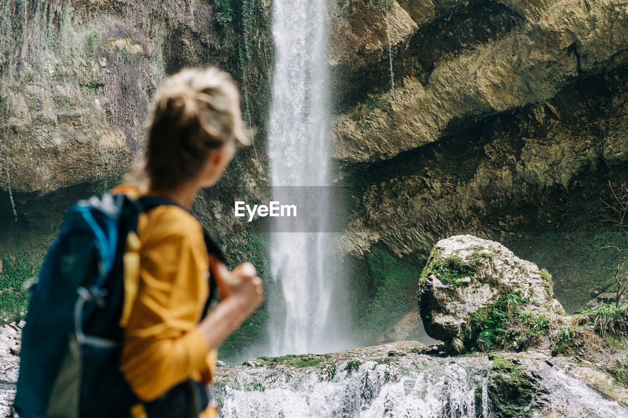 An unrecognizable defocused woman from behind with a backpack stands near a waterfall in a canyon.