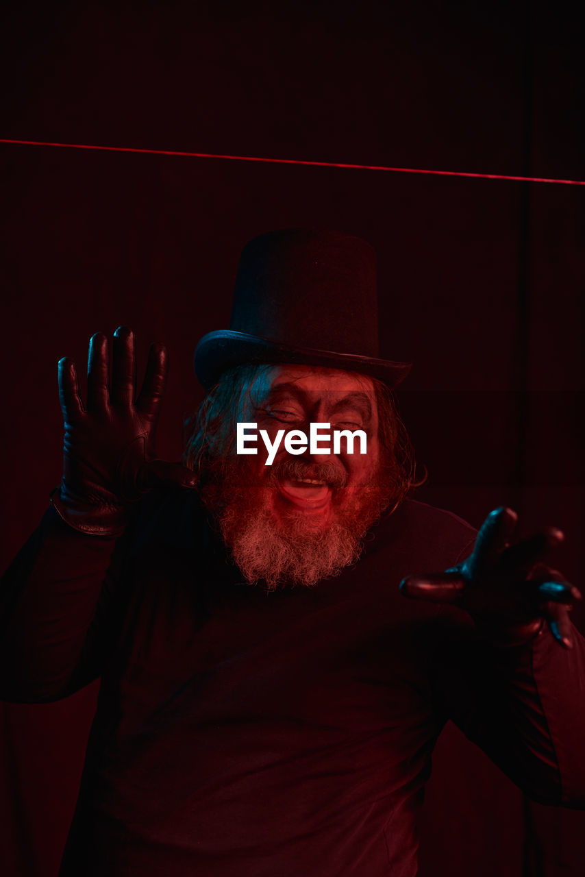 adult, one person, facial hair, hat, portrait, red, men, beard, senior adult, emotion, clothing, performing arts, darkness, fashion accessory, waist up, person, indoors, fedora, stage, looking at camera, human face, entertainment, front view, dark, performance, smiling, studio shot, night, happiness, hand, gesturing