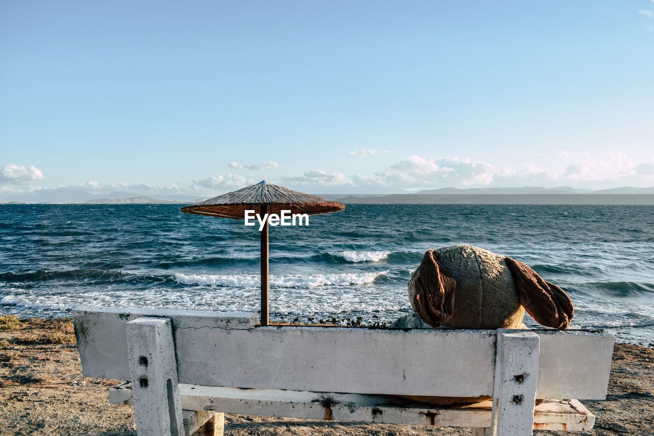 Teddy bear on bench at beach against blue sky during sunny day