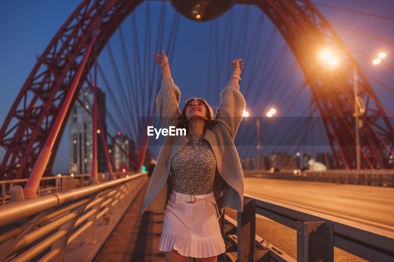 Cheerful woman standing with arms raised on bridge in city at night