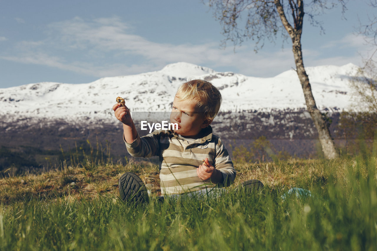 Cute boy sitting on grassy land against snowcapped mountain