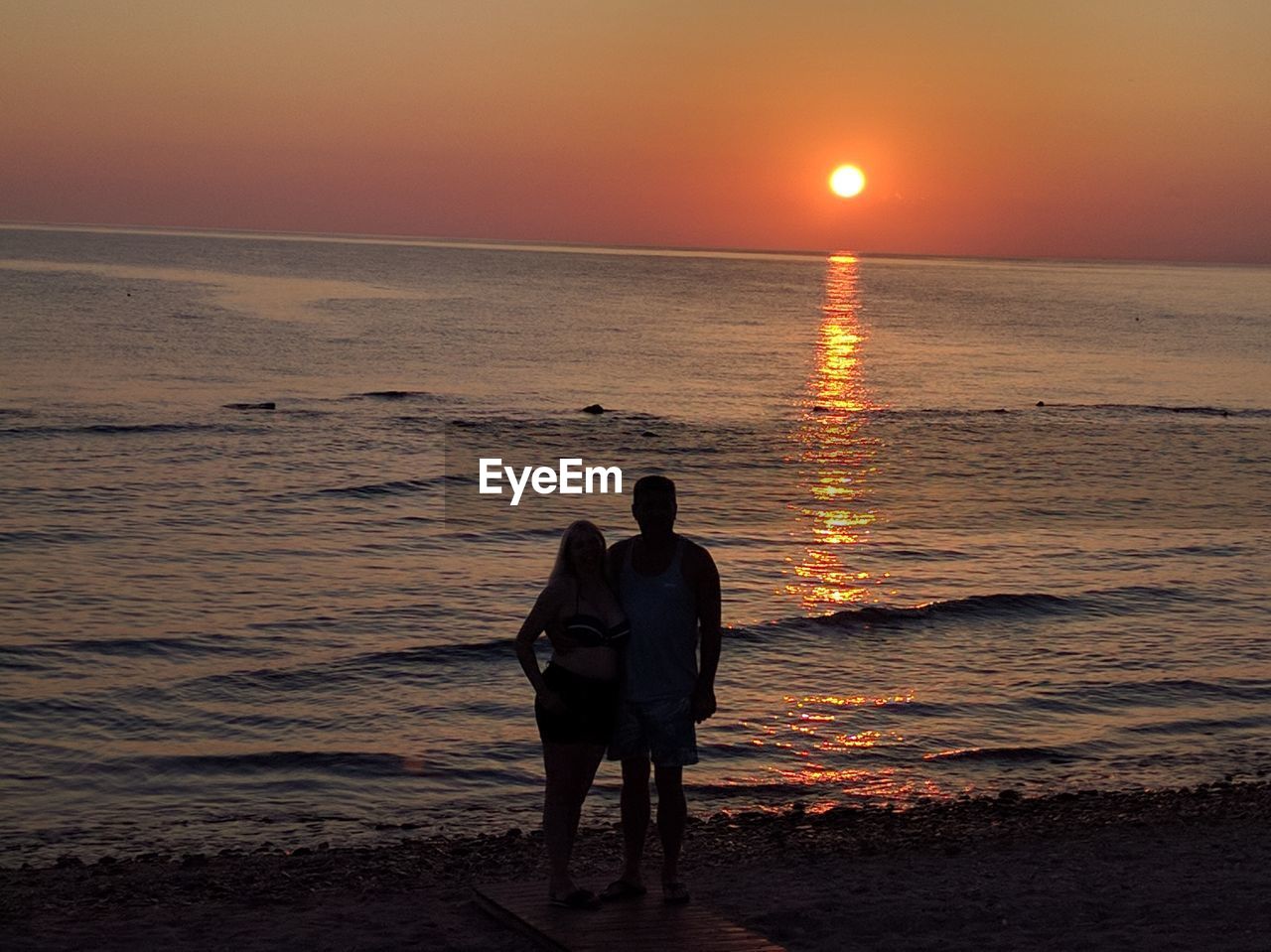 Full length of man and woman standing at beach against sky during sunset