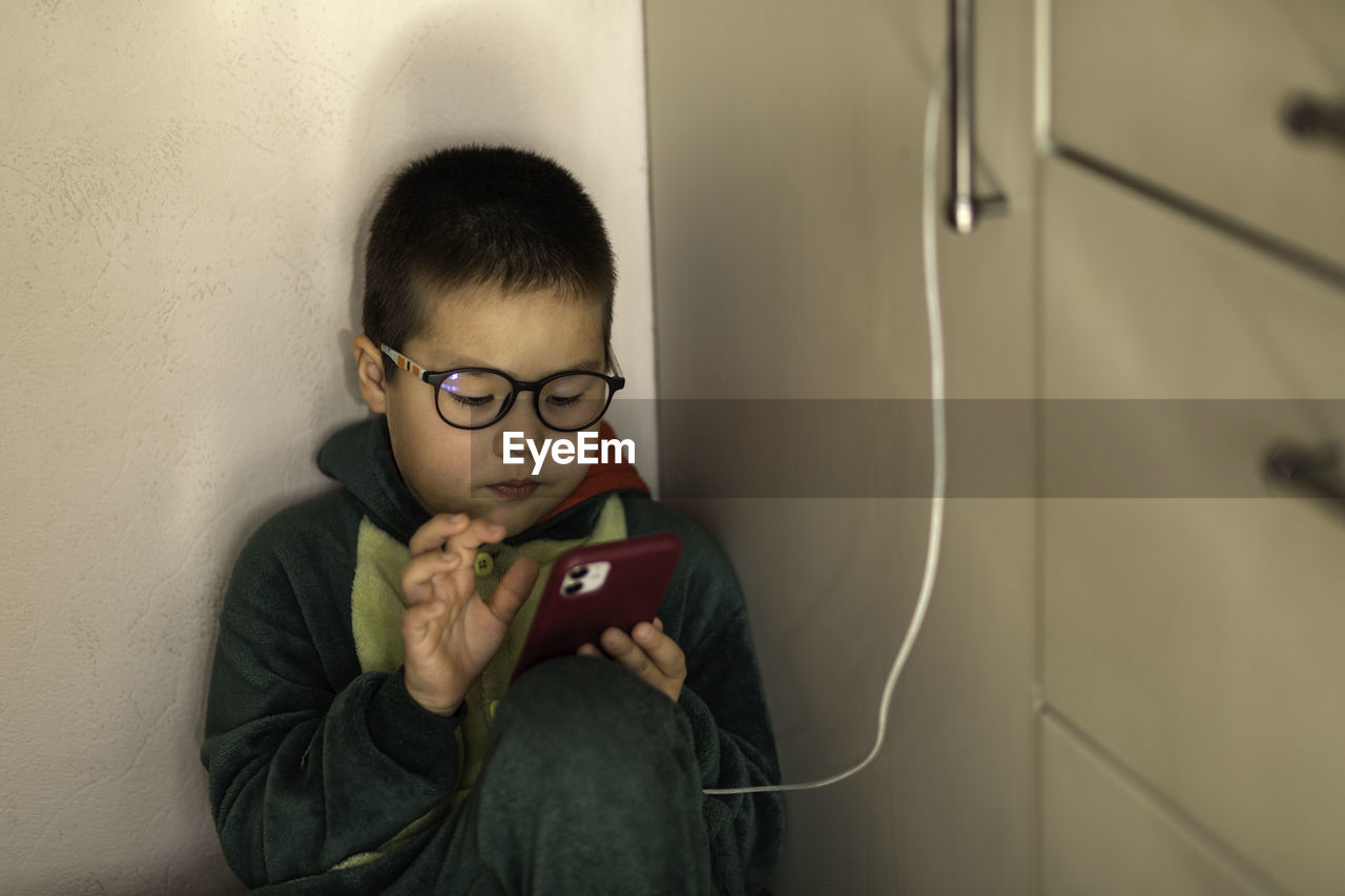 Closeup portrait of a boy who looks into the phone sitting on the floor, phone charging