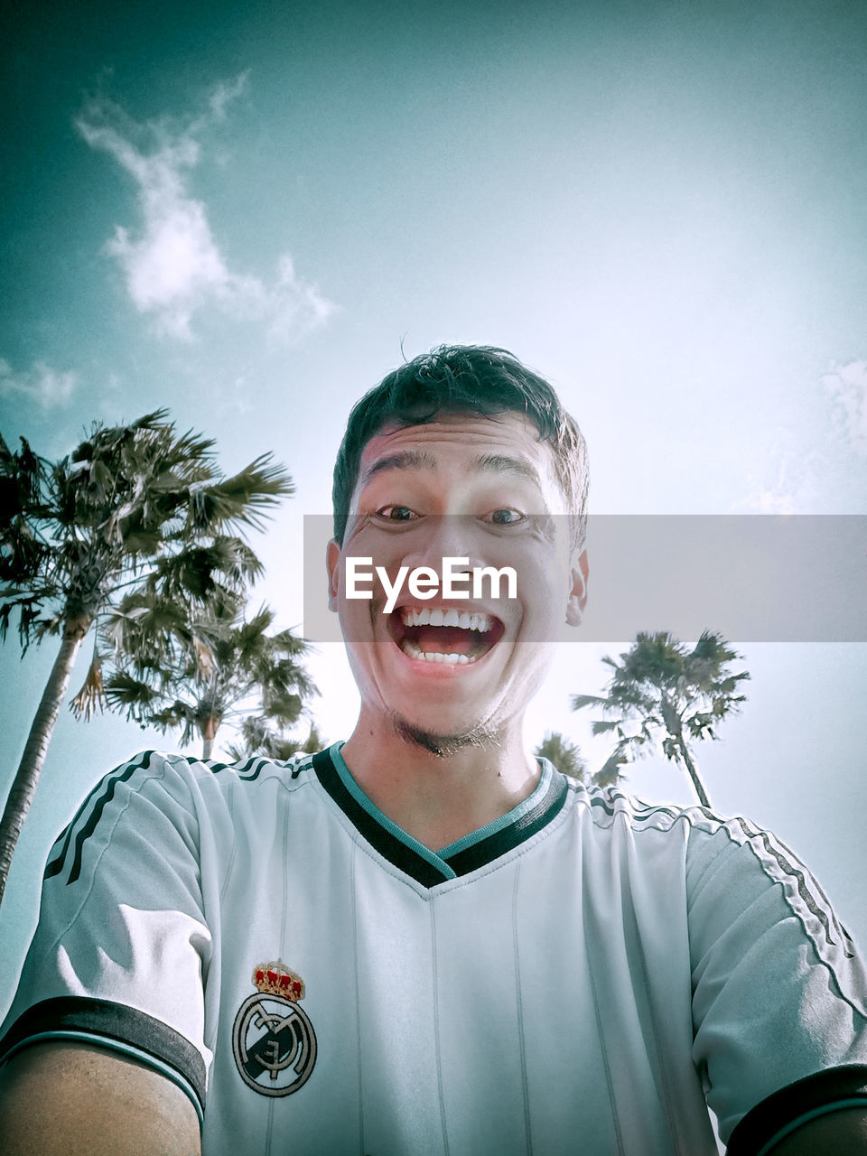 portrait, one person, happiness, sports, smiling, emotion, sky, palm tree, adult, tree, blue, front view, nature, team sport, athlete, young adult, person, soccer, looking at camera, men, clothing, plant, cheerful, cloud, soccer player, waist up, tropical climate, sports uniform, success, outdoors, standing, sports clothing, player, day