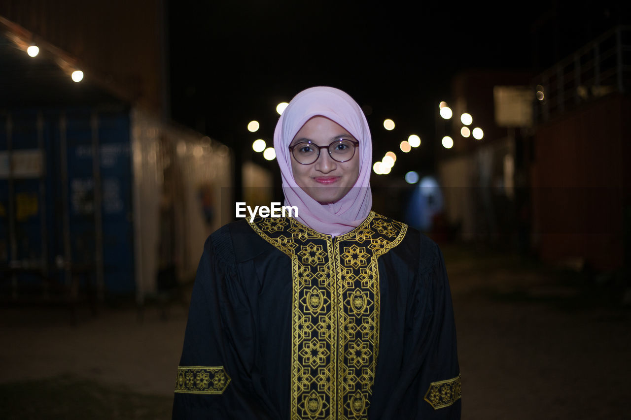 Smiling young woman wearing mortar board while standing against sky at night