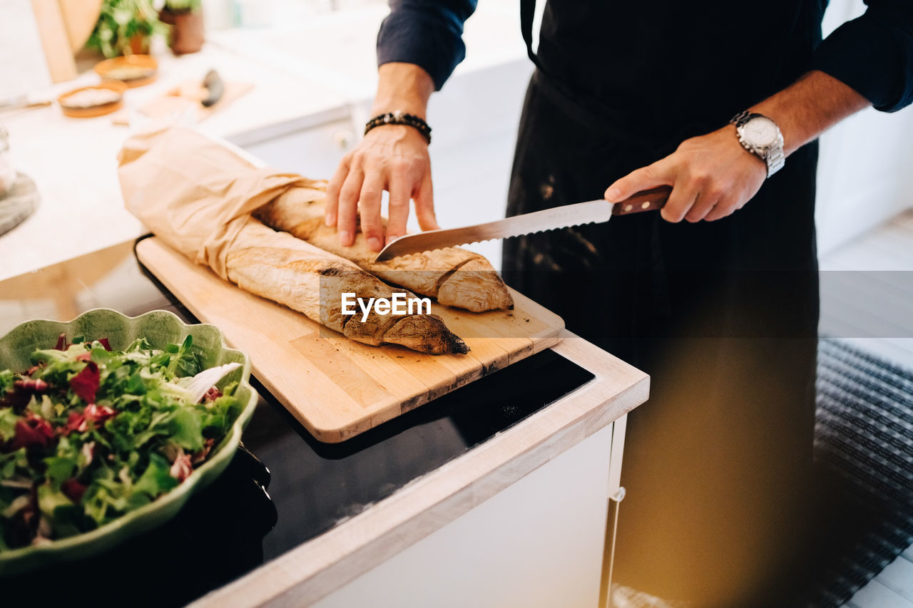 Midsection of man cutting bread by salad at kitchen counter