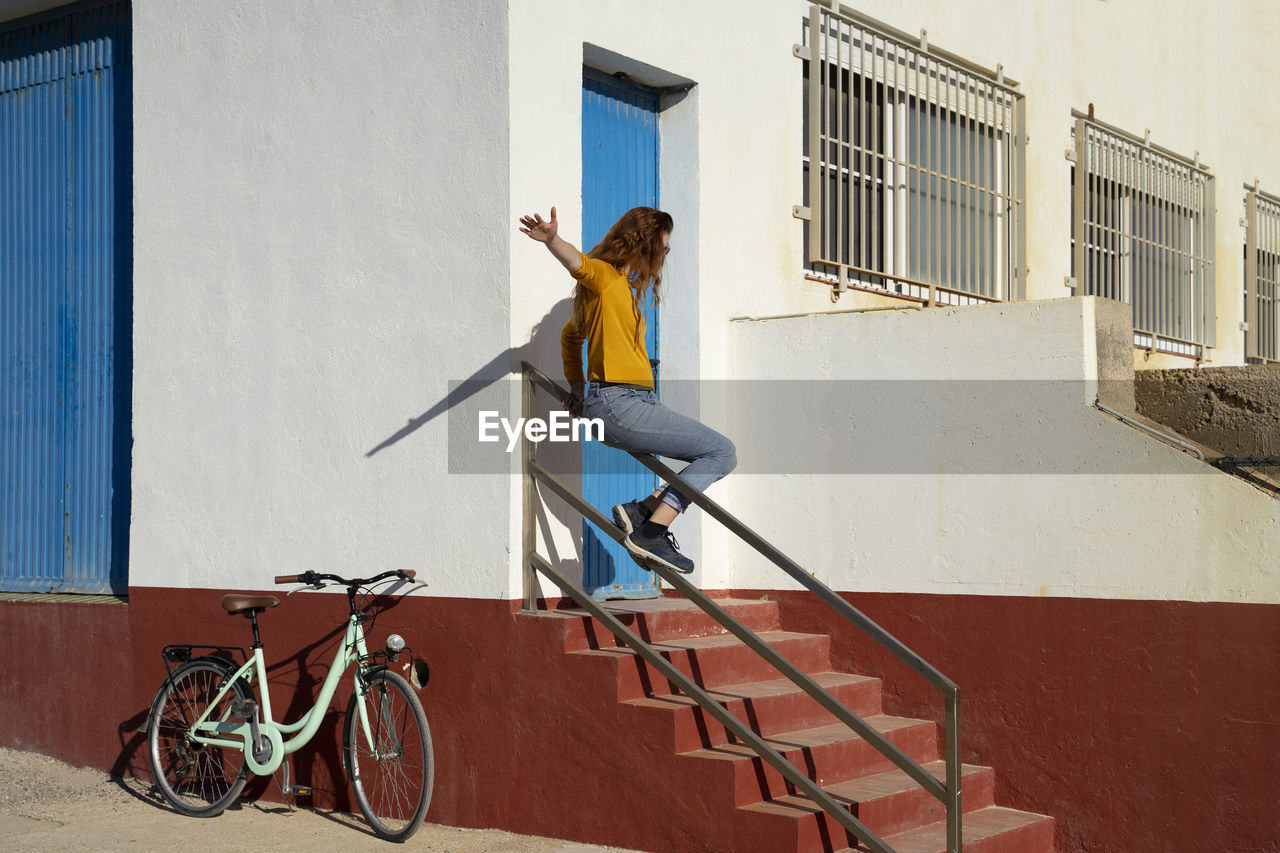 Young woman sliding on railing by white built structure during sunny day