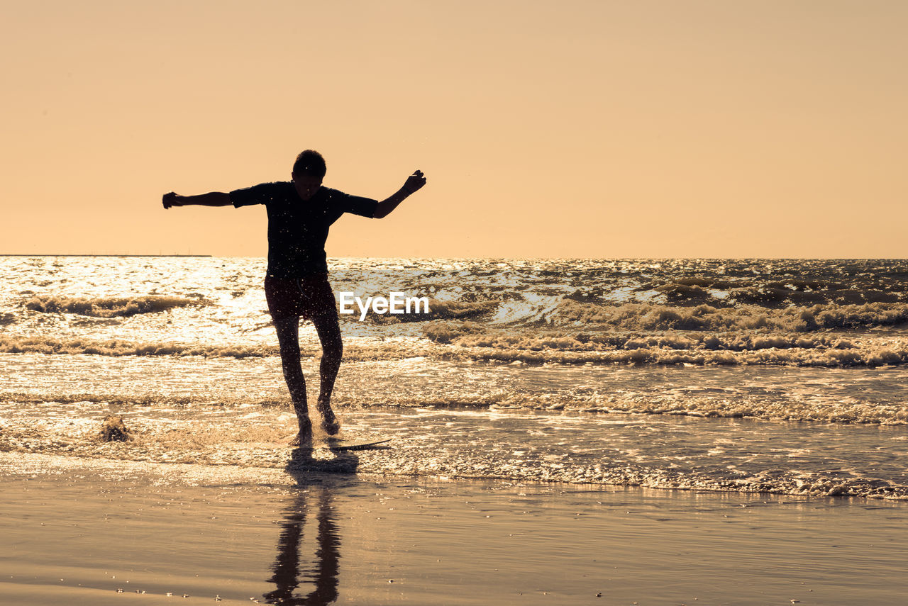 Silhouette young man with arms outstretched surfing on sea against clear sky during sunset