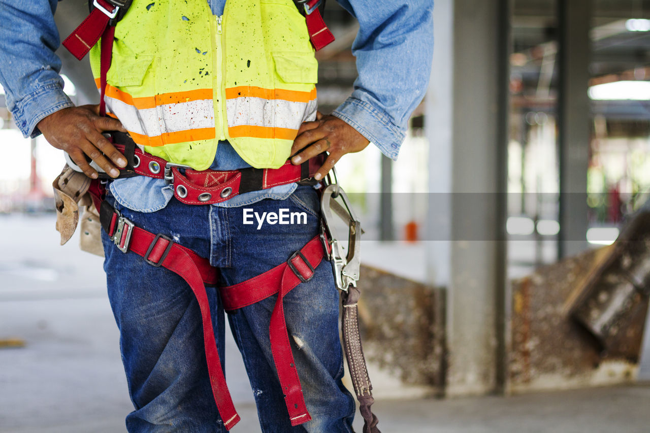 Midsection of manual worker wearing safety harness standing at construction site