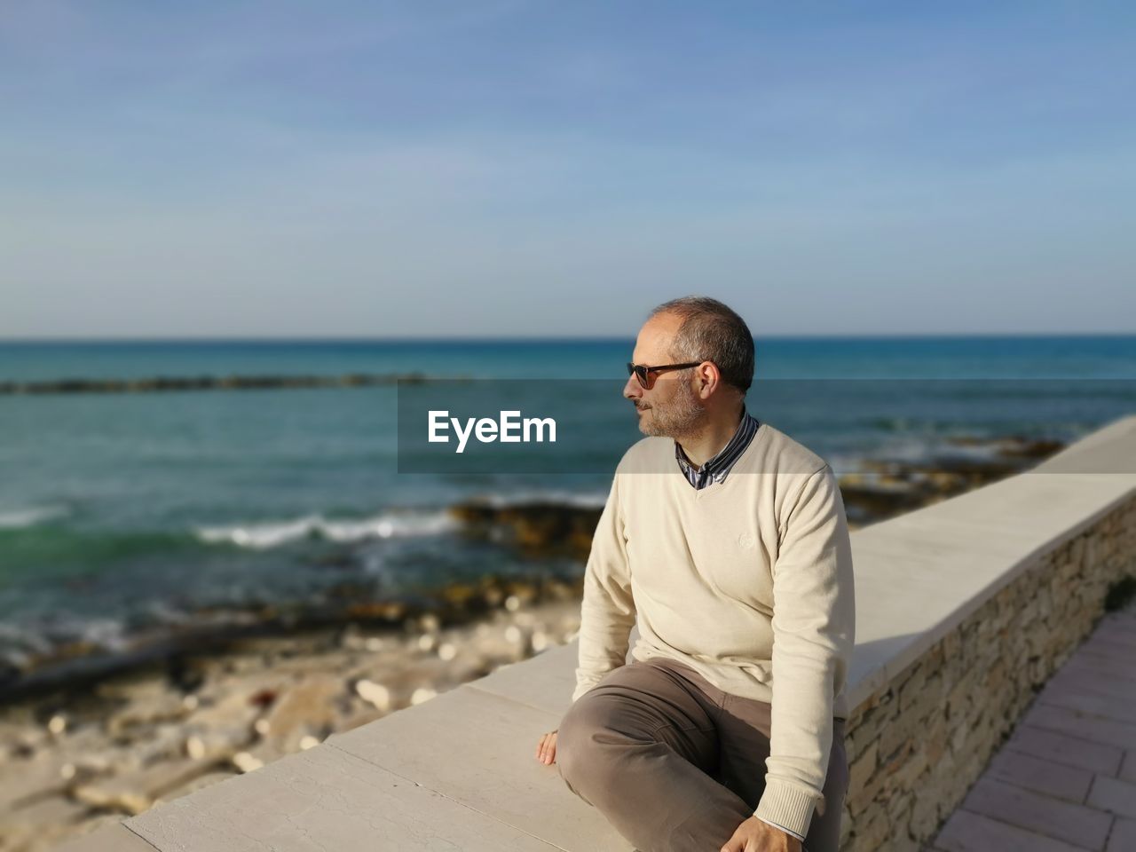 Man looking at sea while sitting on retaining wall against sky