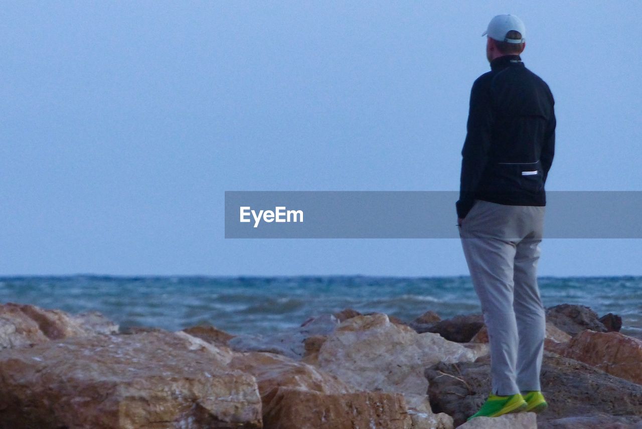 REAR VIEW OF MAN STANDING ON ROCK AT BEACH AGAINST SKY