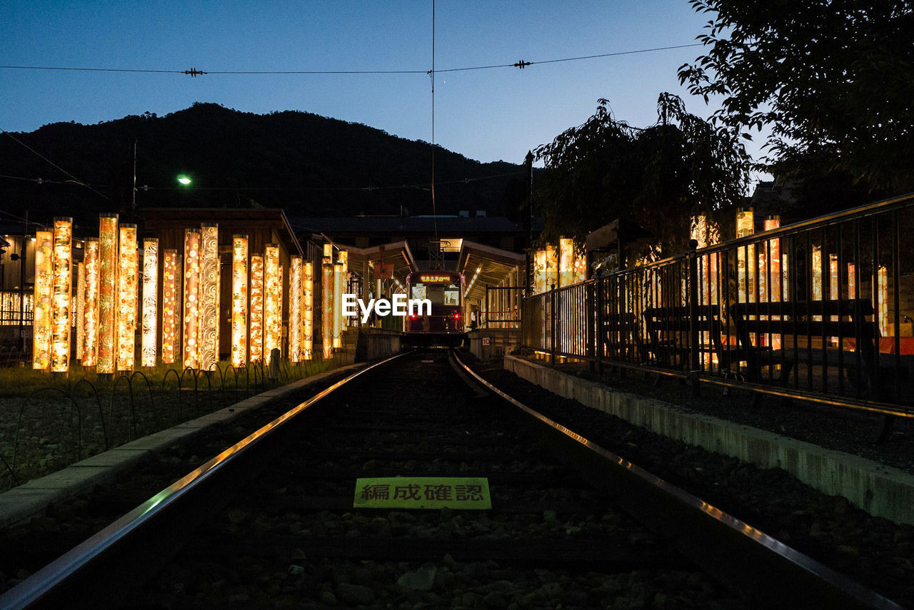 Illuminated railroad station by trees against sky