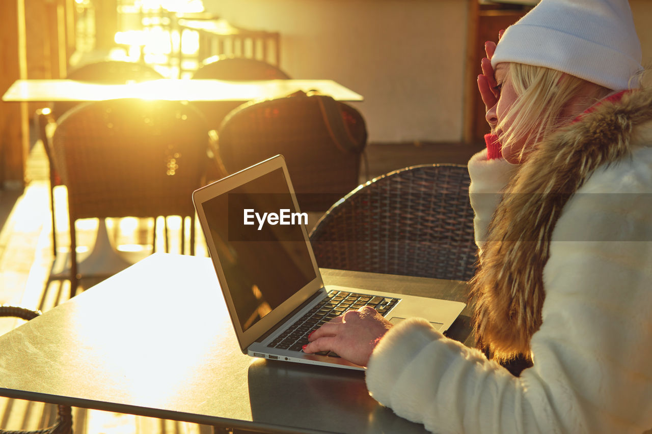 A woman in a sweater and a fur coat is sitting on the terrace of a cafe with a laptop.