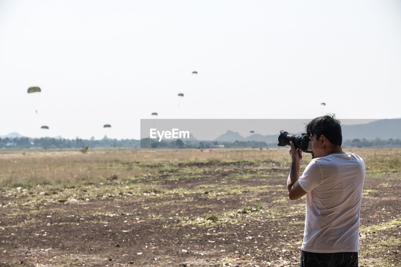 Side view of man photographing on field against cloudy sky