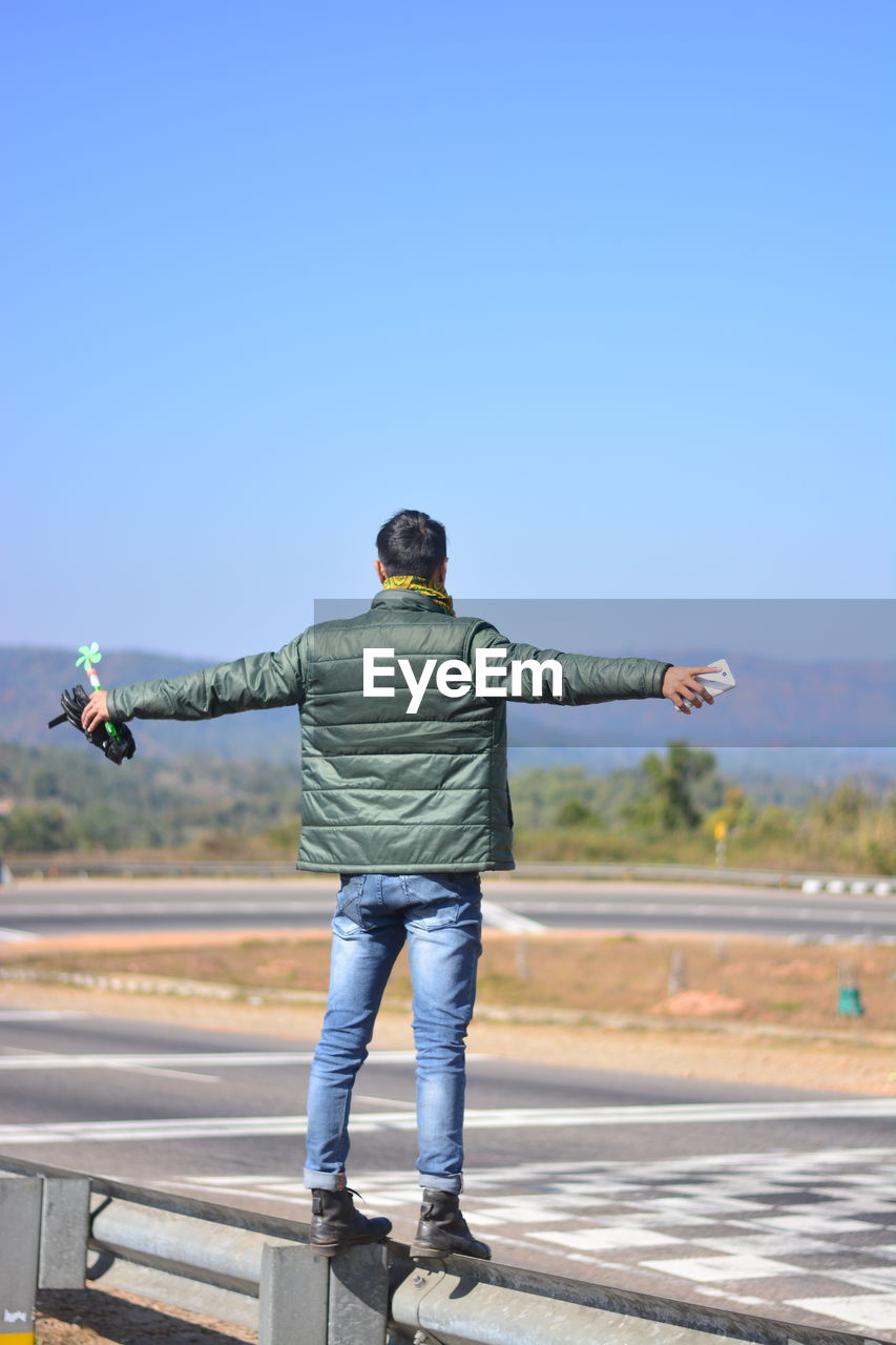 Rear view of man standing on railing against blue sky