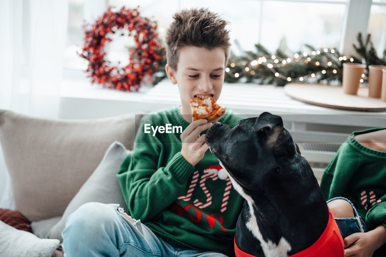 Boy eating pizza while sitting with dog on sofa