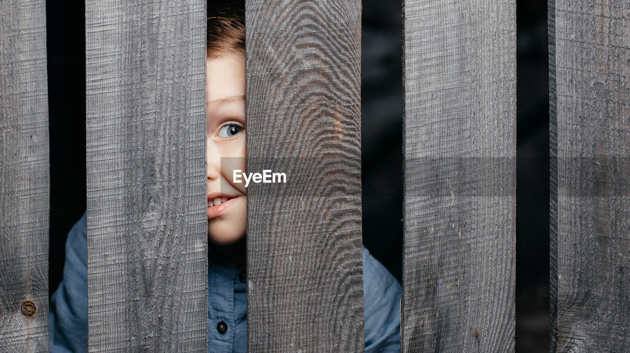 Happy smiling white boy looks out of the crack of a wooden fence. childish curiosity. 