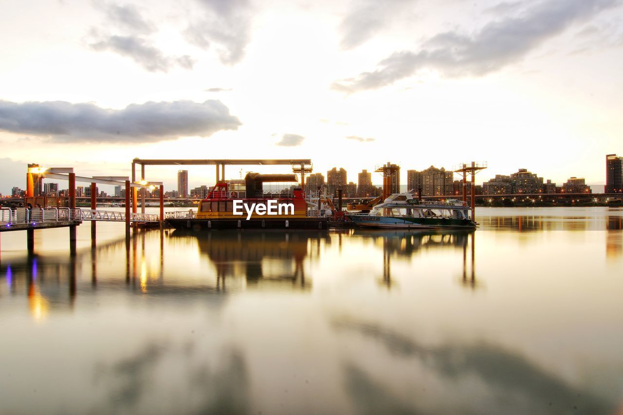 Pier on river by buildings against sky during sunset