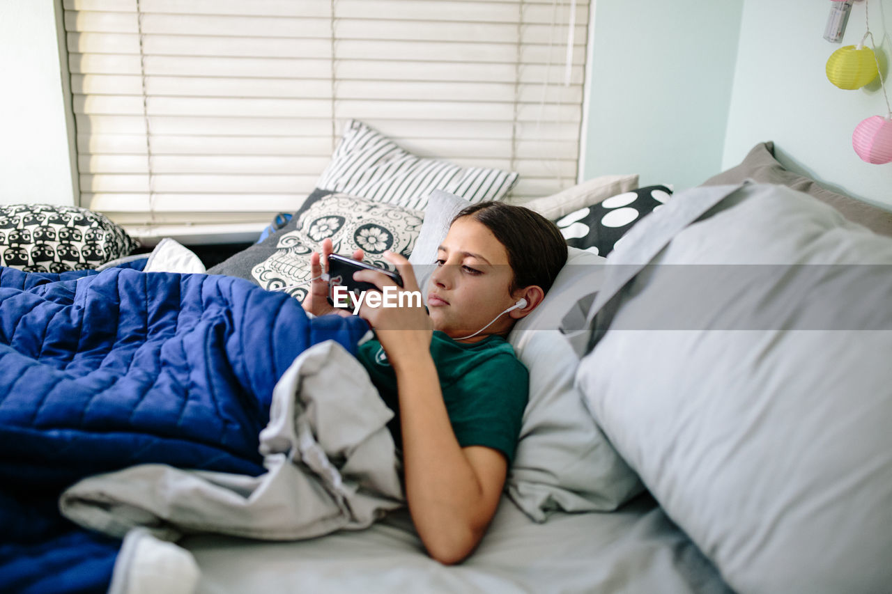 Tween girl lays in bed with her headphones looking at her cellphone