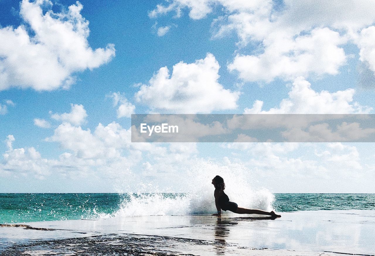Water splashing on silhouette woman posing at beach against cloudy sky
