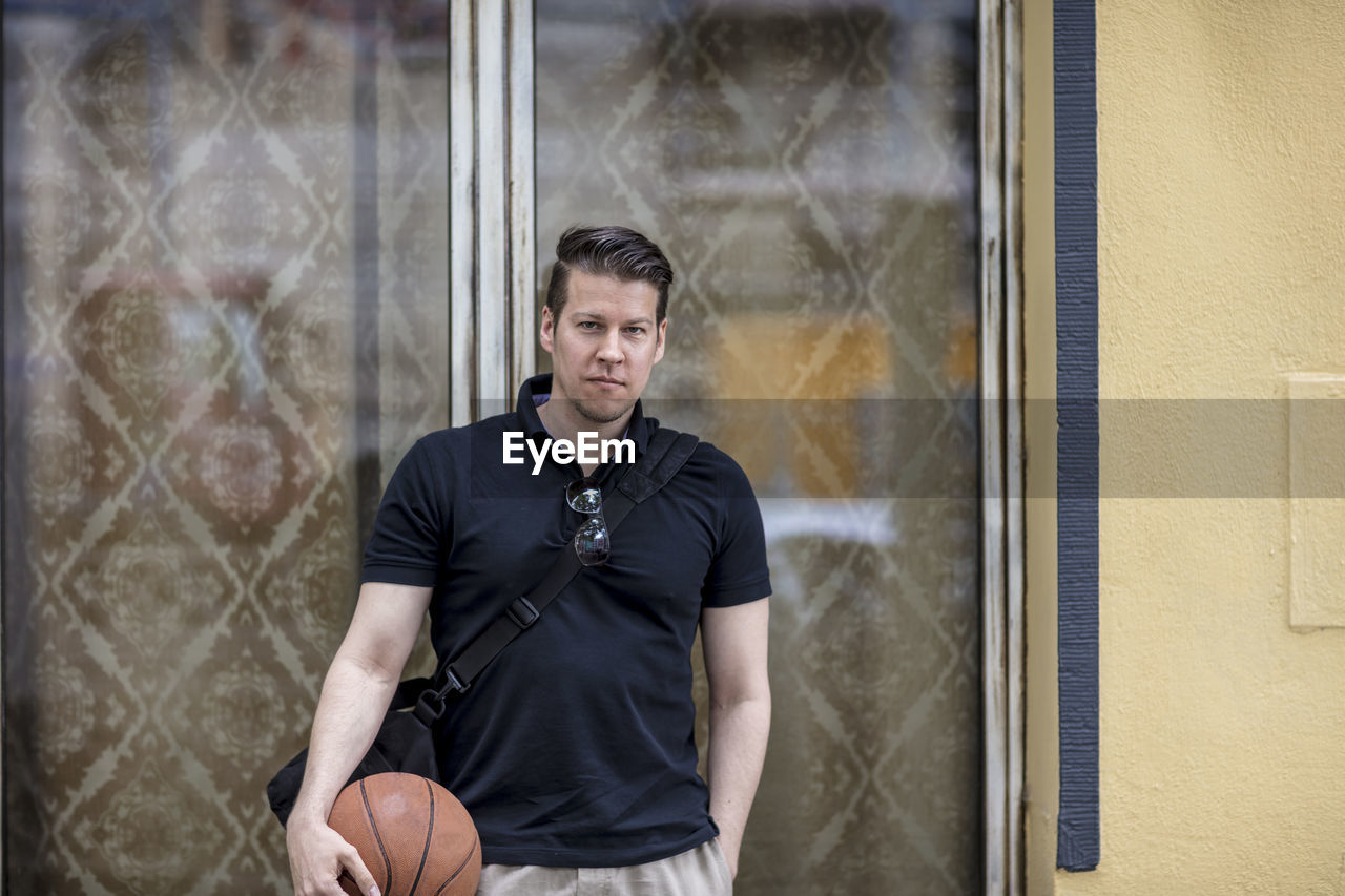 Close-up of mature man holding basketball while leaning against wall