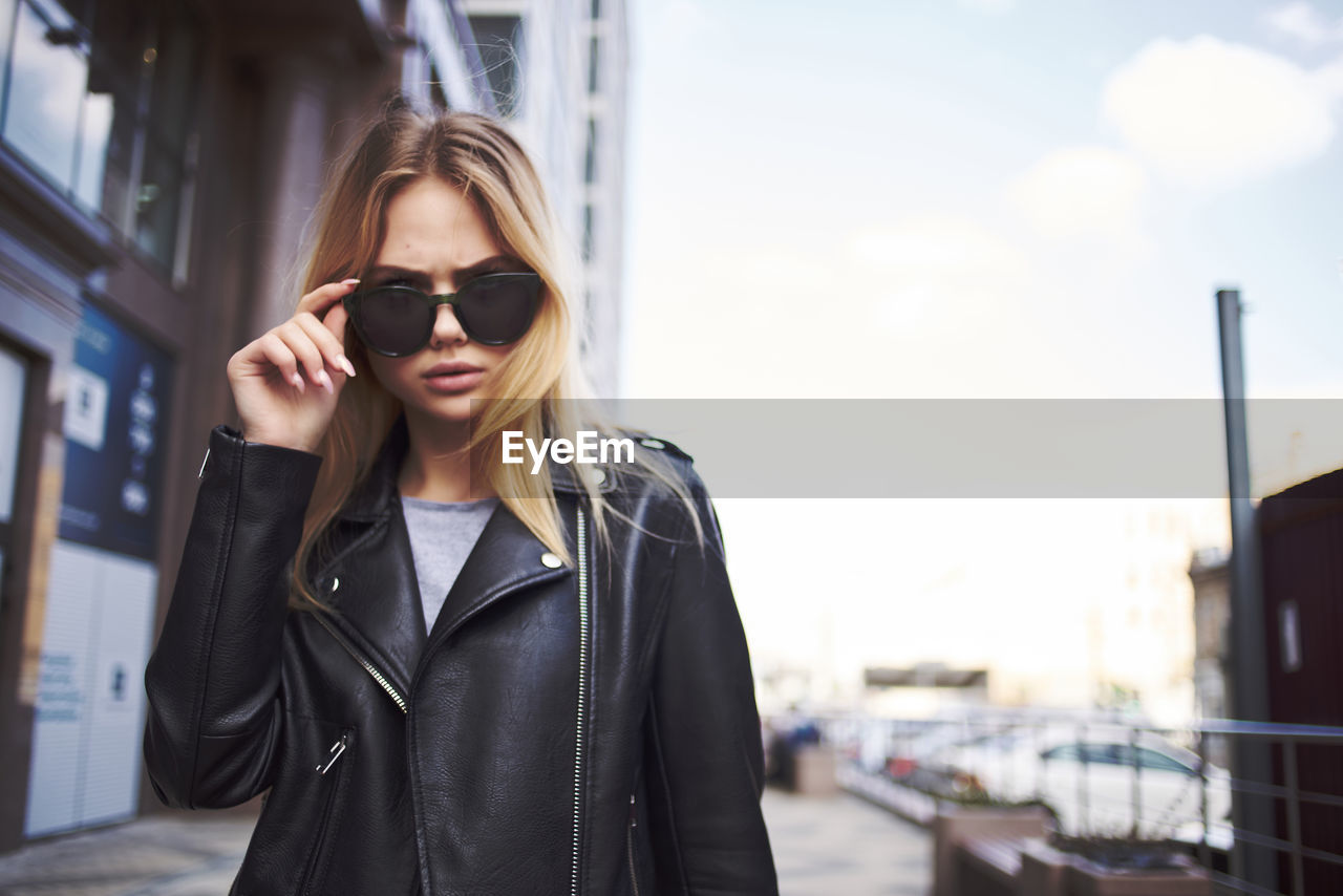 PORTRAIT OF YOUNG WOMAN WEARING SUNGLASSES STANDING AGAINST CAMERA