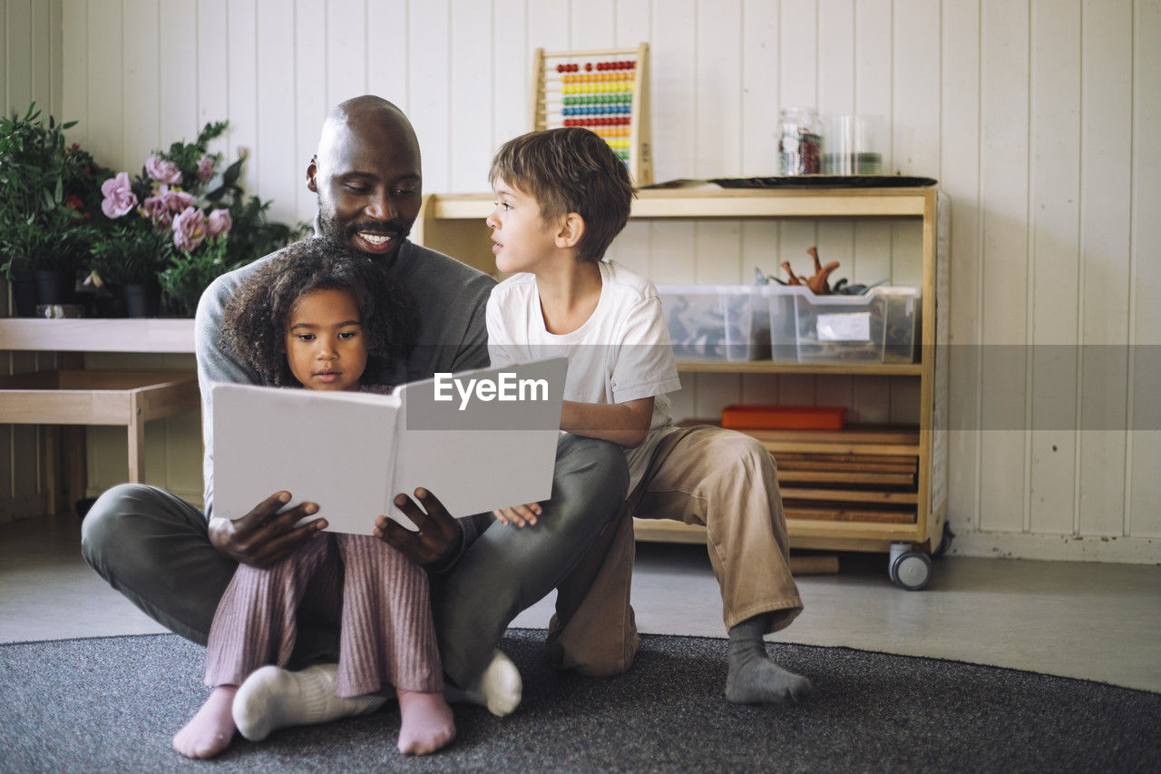 Boy and girl sitting with male teacher holding book while sitting cross-legged in classroom at kindergarten