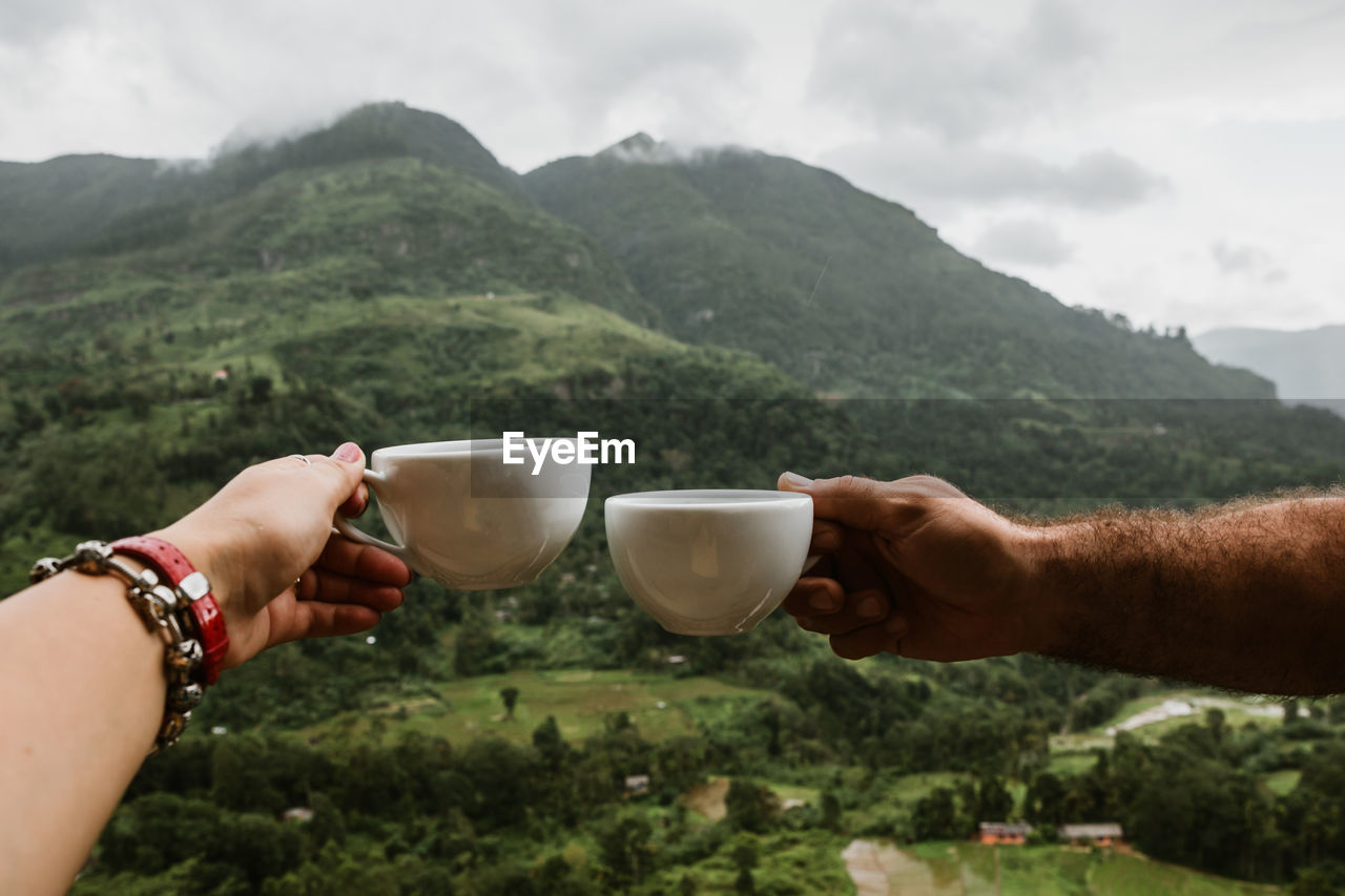 Cropped hands of couple toasting coffee cups against mountain