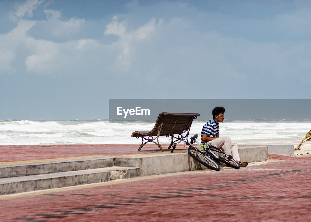 Young man with bicycle sitting on promenade against sky