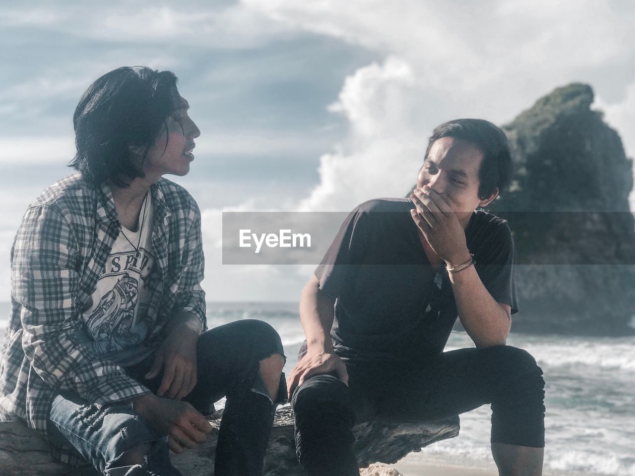 Young men sitting on rock at beach against sky