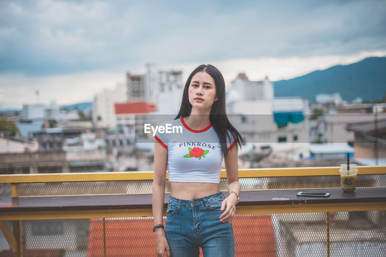 Portrait of young woman standing against cloudy sky in city