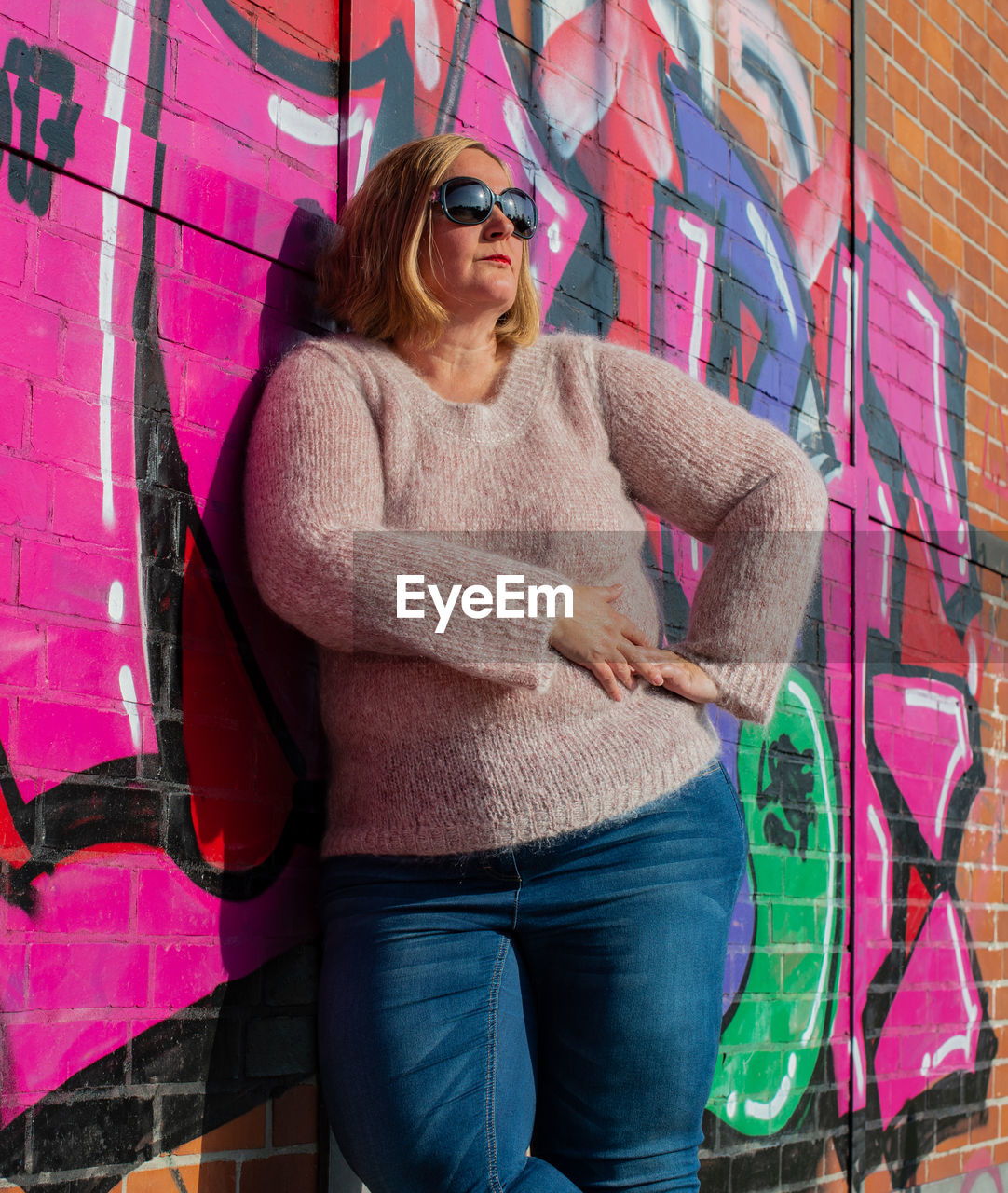 YOUNG WOMAN WEARING SUNGLASSES STANDING AGAINST GRAFFITI WALL