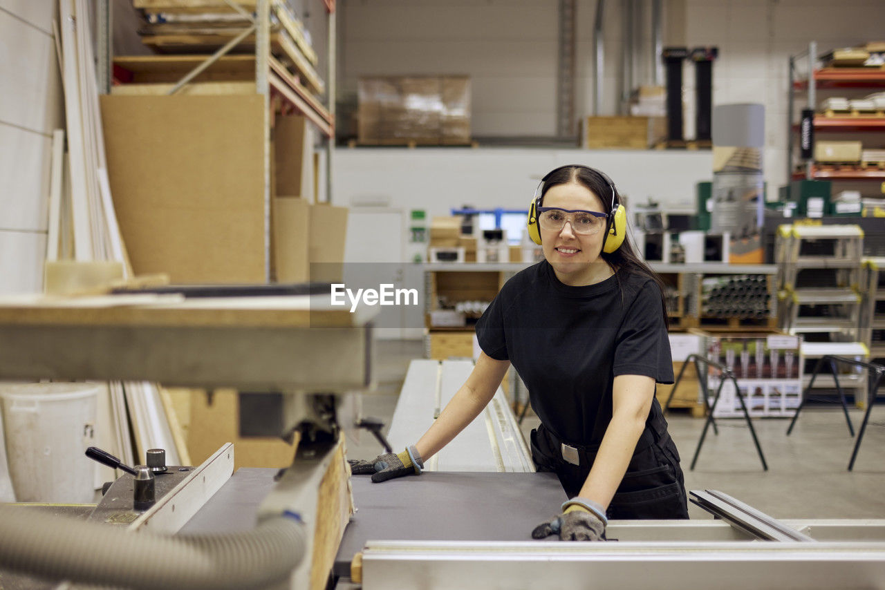 Portrait of smiling female carpenter wearing protective eyewear and ear protectors while working in industry
