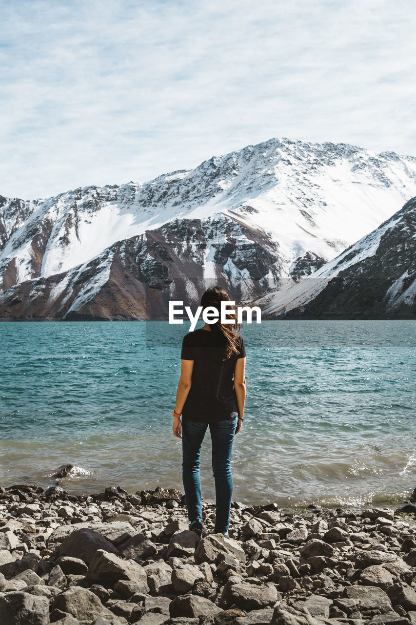 Rear view full length of woman standing at lakeshore against snowcapped mountain