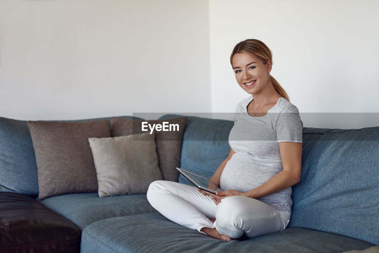 Portrait of smiling pregnant young woman with digital tablet sitting on sofa at home