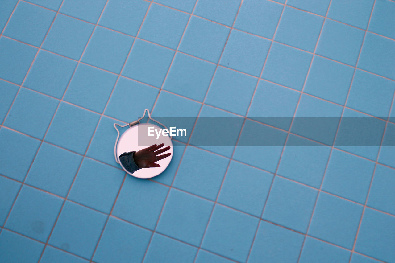 High angle view of hand reflection on mirror over blue tiled floor