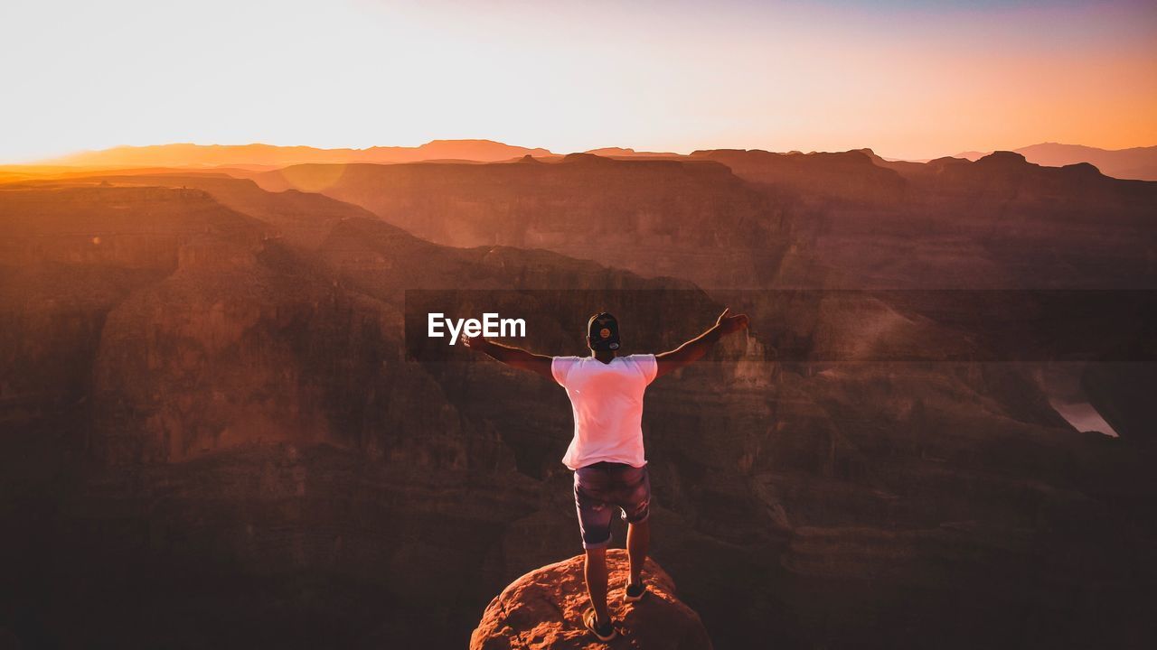 REAR VIEW OF MAN STANDING ON MOUNTAIN LANDSCAPE AGAINST SKY