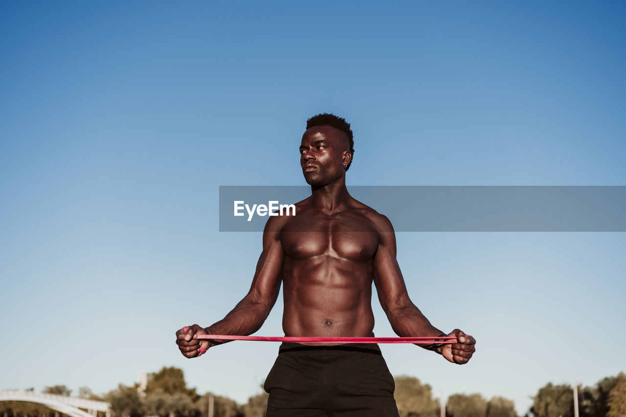 Male athlete looking away while stretching resistance band standing against clear sky during sunset