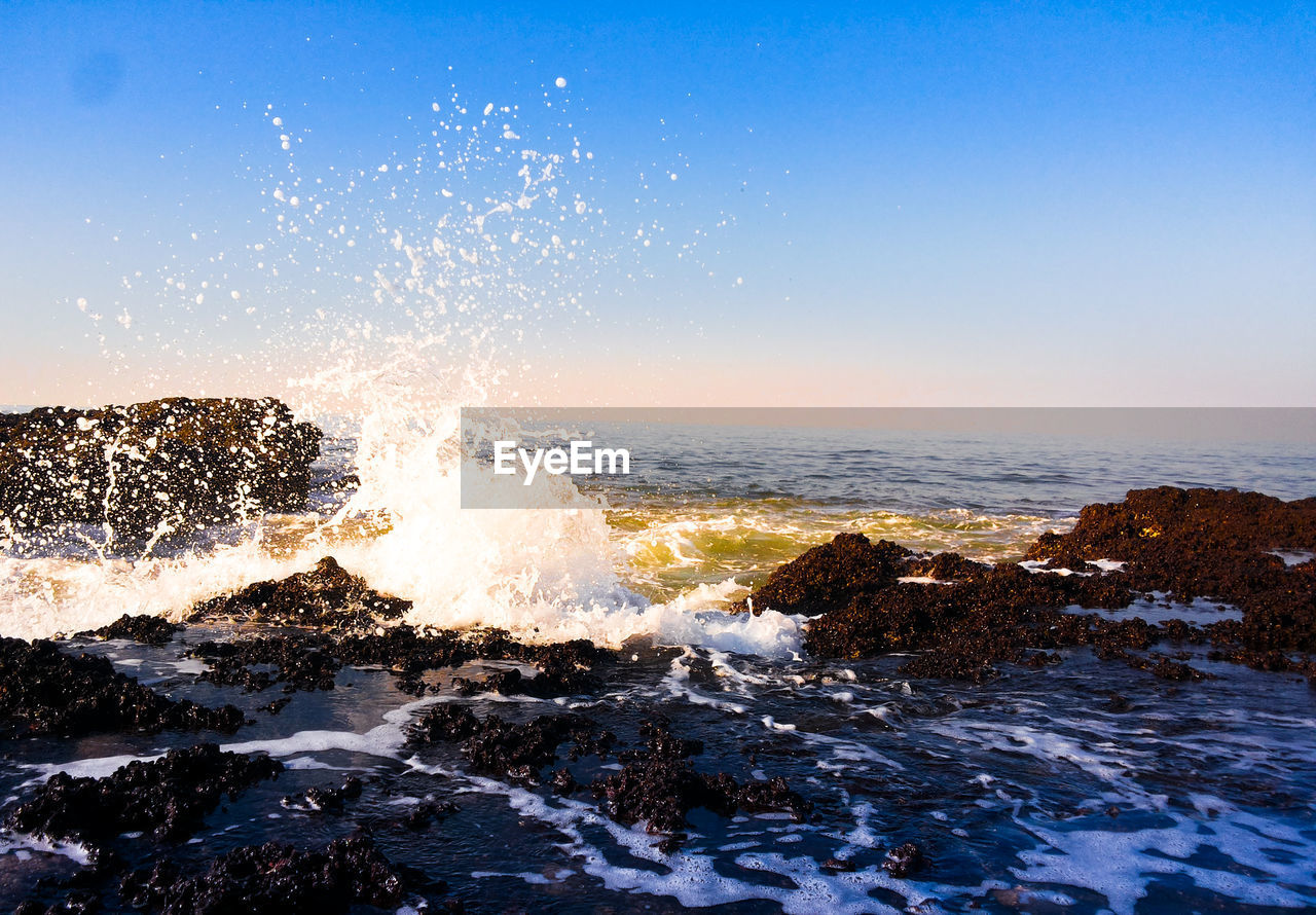 Waves splashing on rocks against clear blue sky