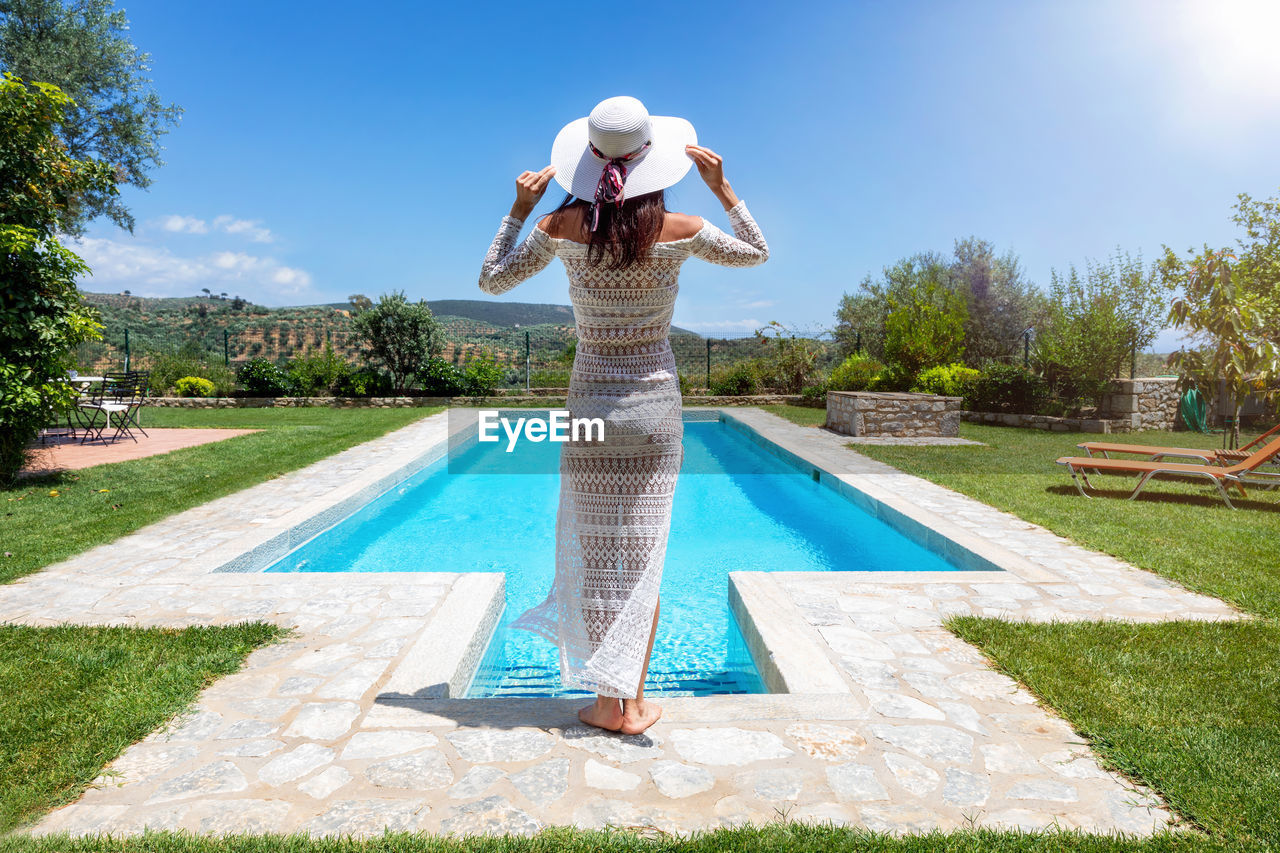 WOMAN STANDING BY SWIMMING POOL IN PARK AGAINST SKY