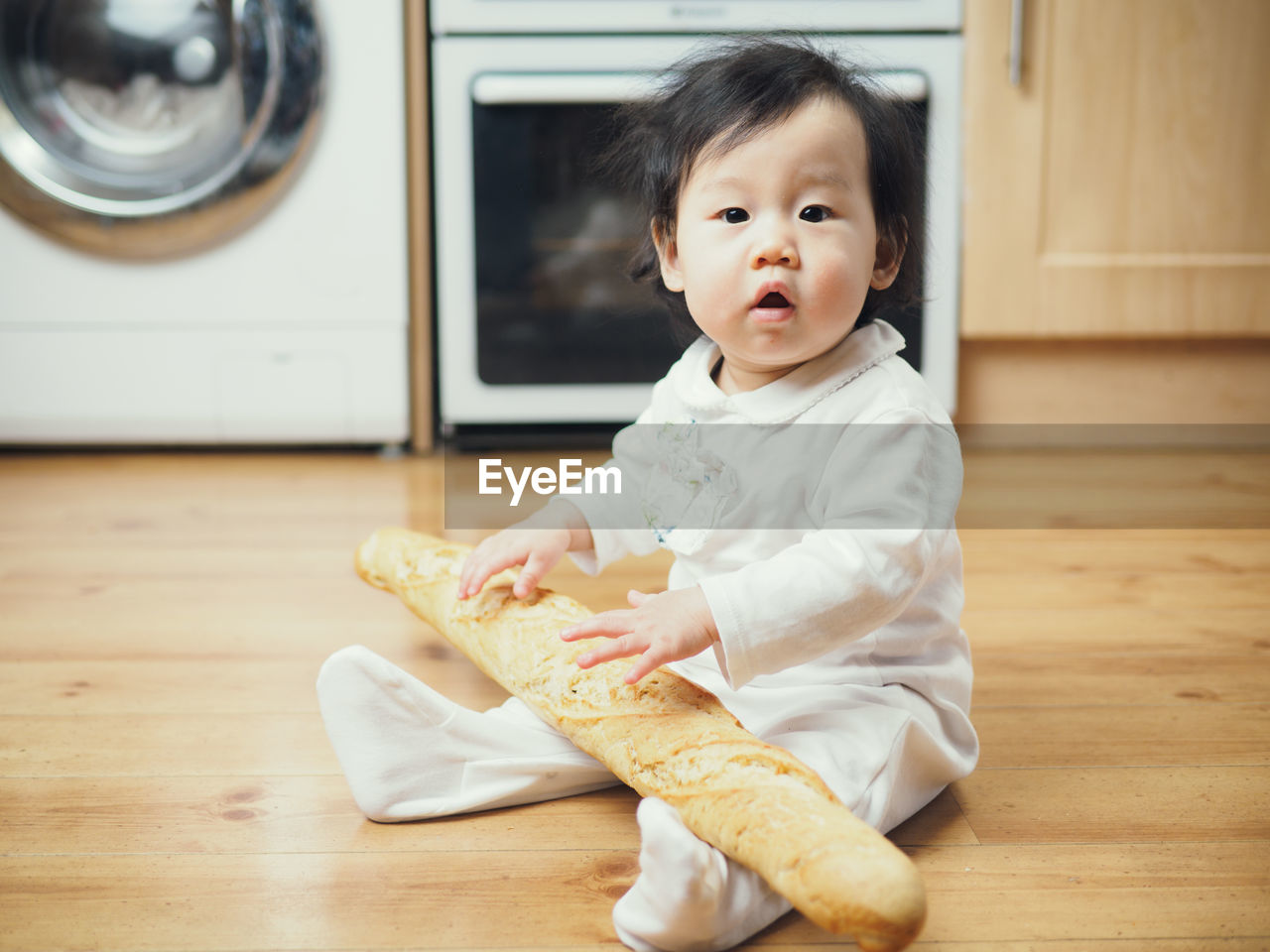 Cute baby girl sitting with baguette on hardwood floor at home