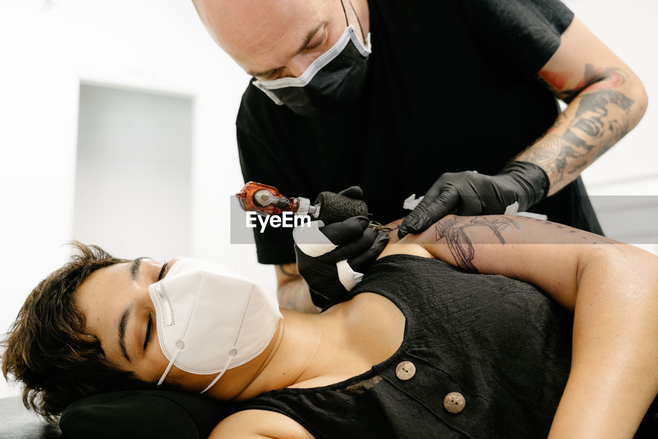 Tattooist with machine making tattoo on arm of woman lying on table in salon